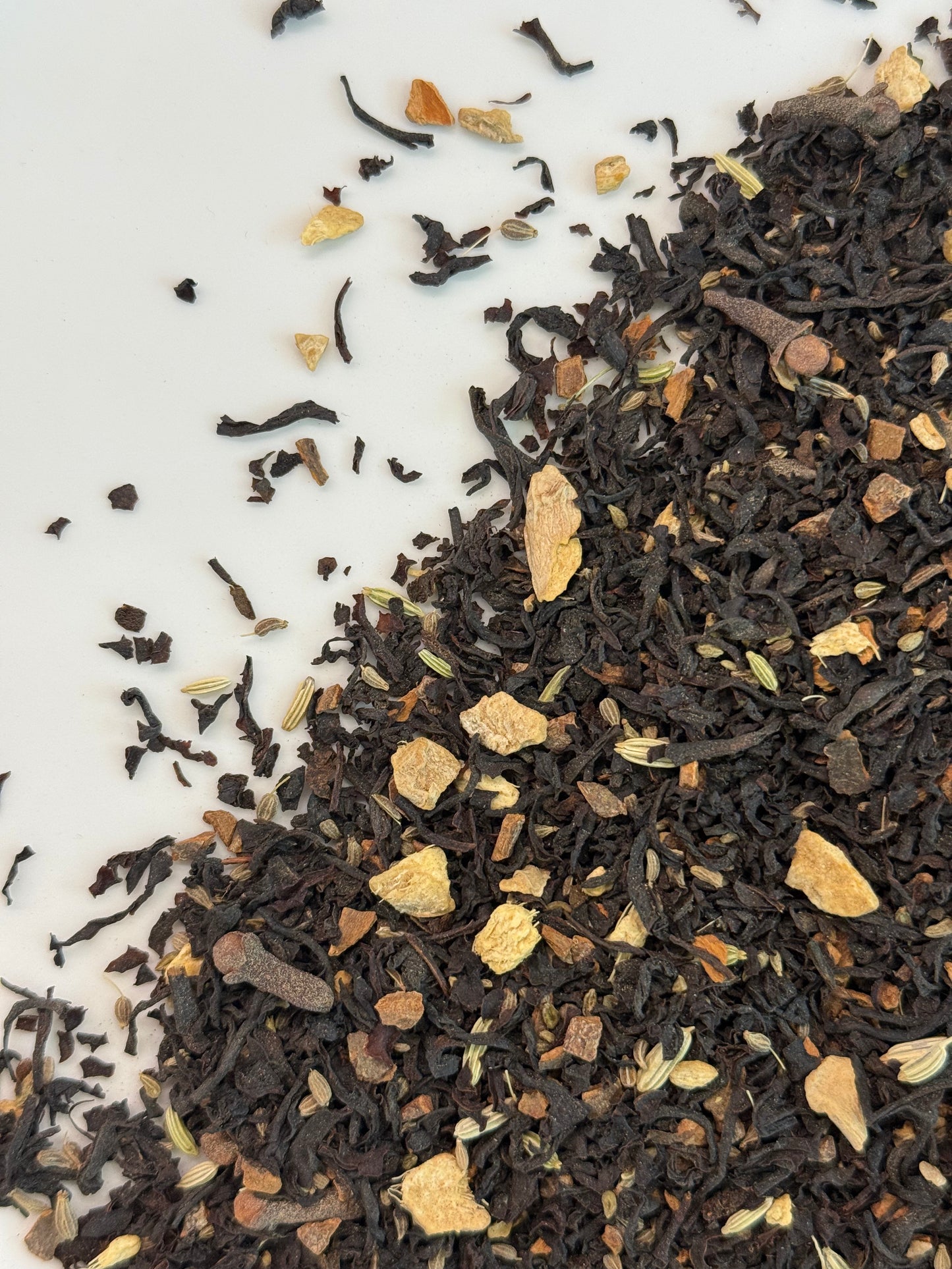 A close-up top-down view of loose-leaf tea blend spilled across a white surface. The tea consists of dark brown to black dried leaves mixed with small tan, beige, and light brown botanical pieces.