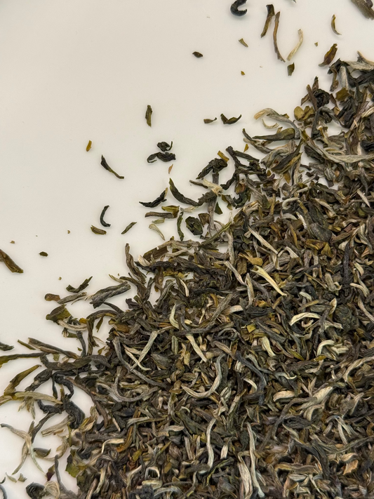 Dried loose-leaf tea leaves scattered diagonally across a plain white surface, showing dark green, olive, and light silver-gray tones.