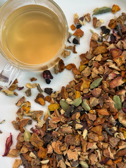 A glass mug filled with pale golden teasine pumpkin chai sits on a white surface beside spilled loose dried botanicals, including orange peel, cardamom pods, and flower pieces.