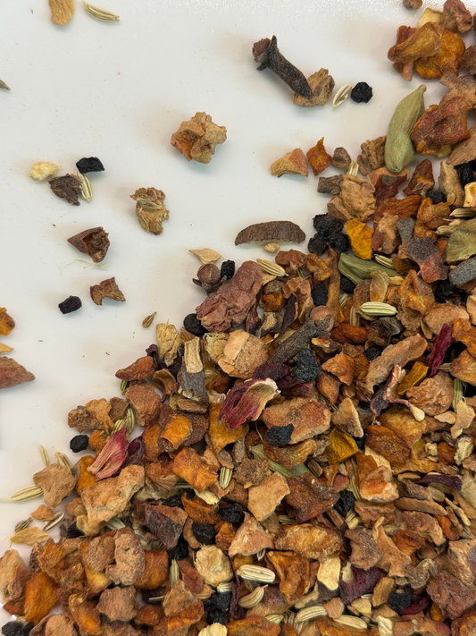 A close-up of loose dried herbal tea ingredients scattered on a white surface, including orange dried flowers, brown bark pieces, small green pods, and dark berries.