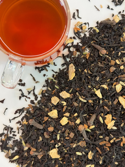 A close-up overhead view of loose-leaf tea, showing dark dried tea leaves mixed with light tan citrus pieces and whole spices, with a glass cup of amber tea in the upper left on a white surface.