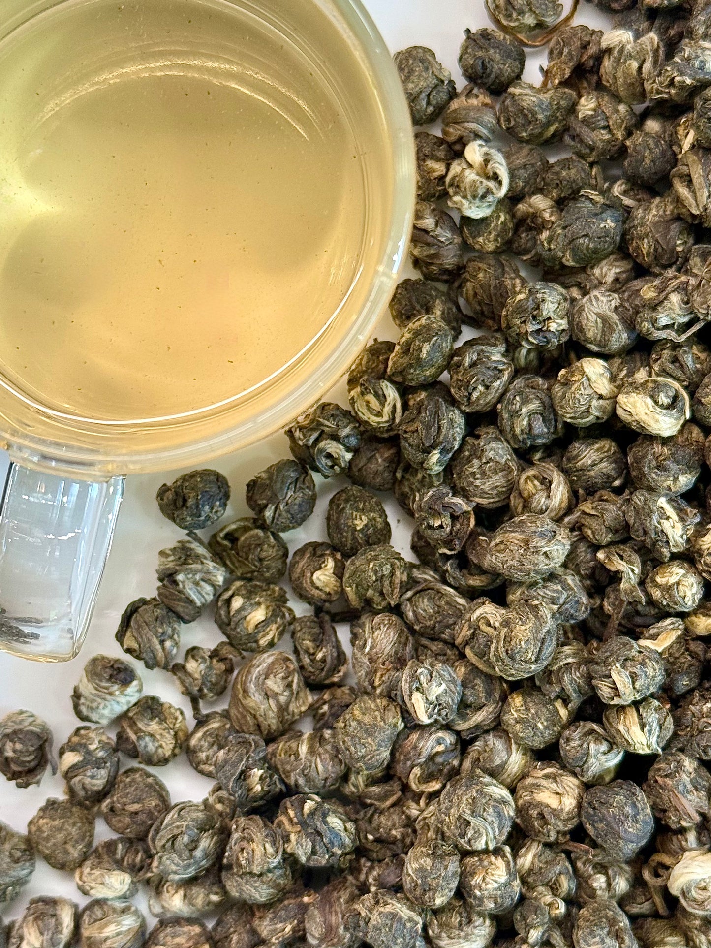 A close-up of loose rolled green tea leaves with a clear glass cup filled with brewed pale yellow tea on a white surface.